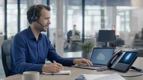 Cloud-Telefonanlage mit Microsoft Teams: Mitarbeiter telefoniert über Headset am Laptop, daneben ein für Teams zertifiziertes Tischtelefon