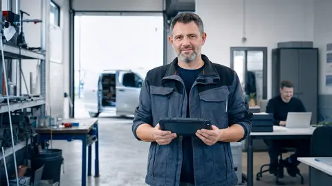 IT für Handwerker: Handwerksmeister mit robustem Tablet in der Werkstatt, im Hintergrund das Büro des Handwerksbetriebs mit Laptop und der Servicewagen vor dem Tor